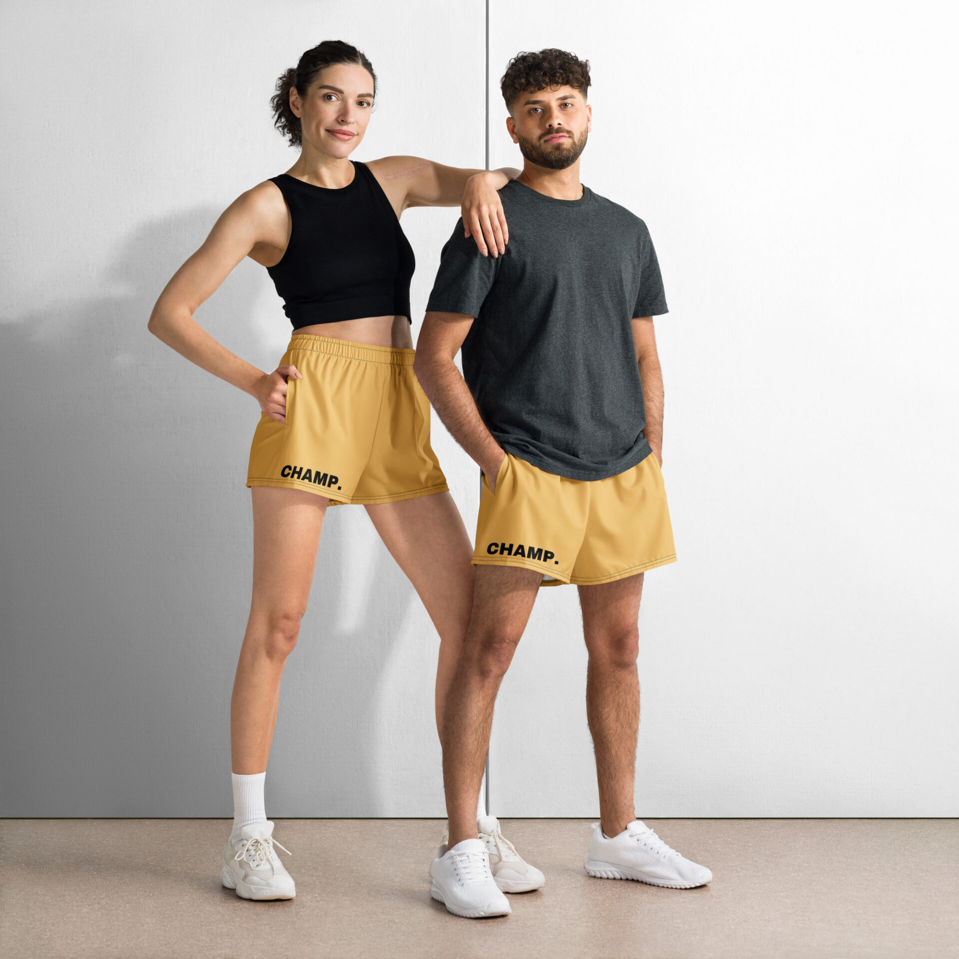 A woman in a black tank top and CHAMP Golden Athletic Shorts stands confidently alongside a man in a gray t-shirt and matching shorts. CHAMP. is emblazoned on their golden shorts, as both have hands in pockets, sporting white sneakers against a two-tone wall.
