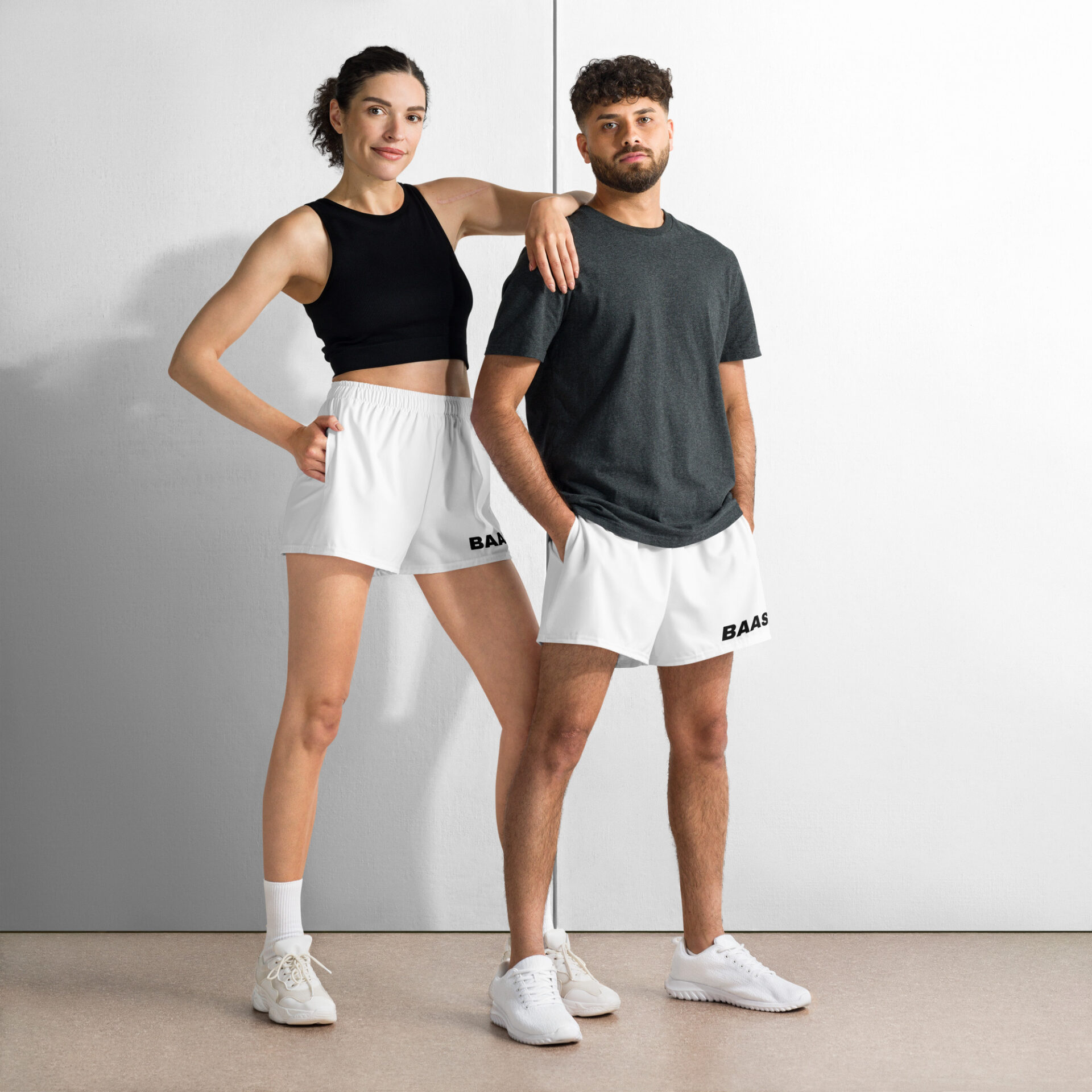 A woman and man stand confidently against a white wall; she in a black sleeveless top and athletic shorts, he in a dark gray T-shirt and BAAS Athletic Shorts. Both complete their look with crisp white sneakers.