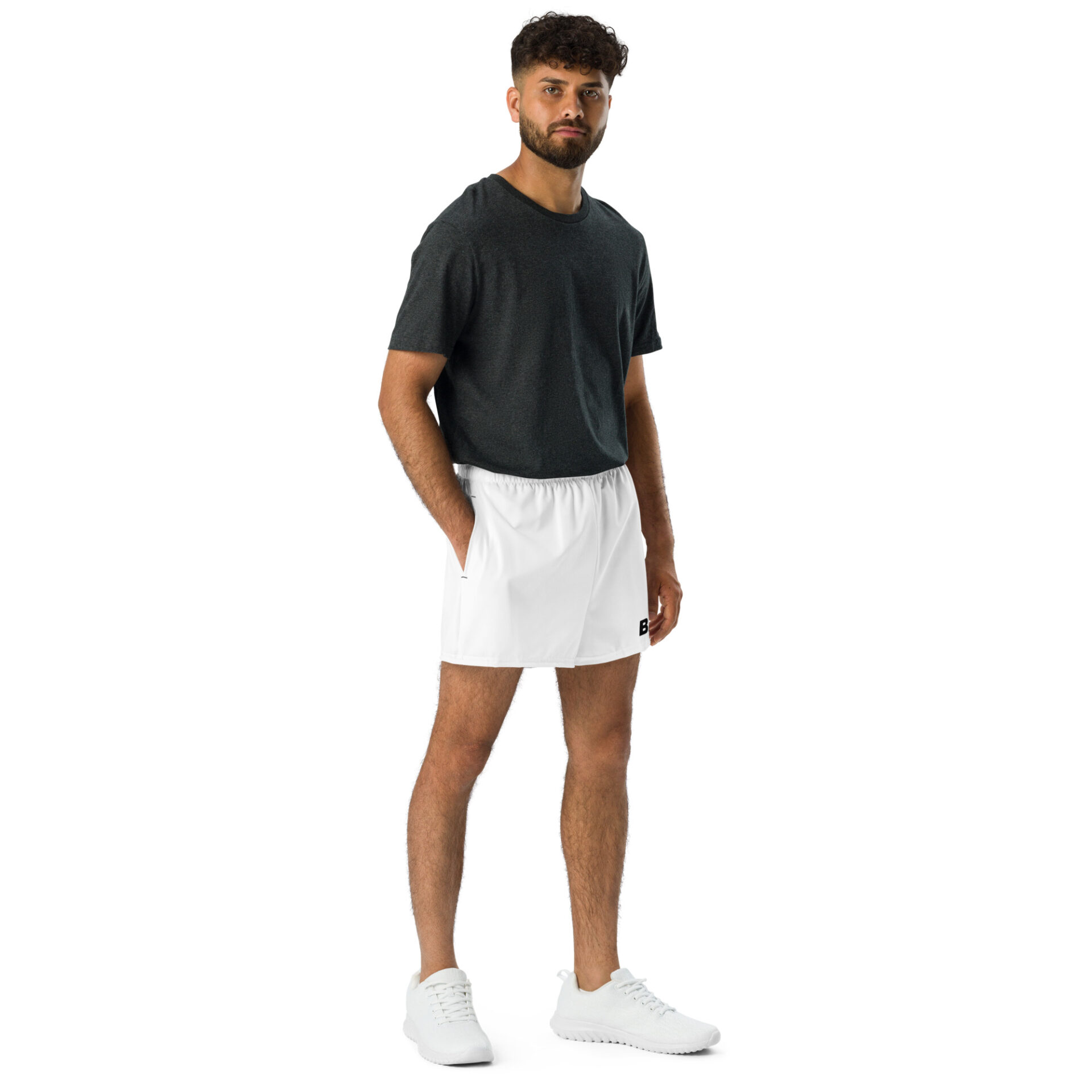 A man poses against a white background, showcasing the latest in sportswear: a dark gray t-shirt, BAAS Athletic Shorts in crisp white, and sleek white sneakers. His relaxed arms, curly hair, and beard complete the effortless BAAS look.