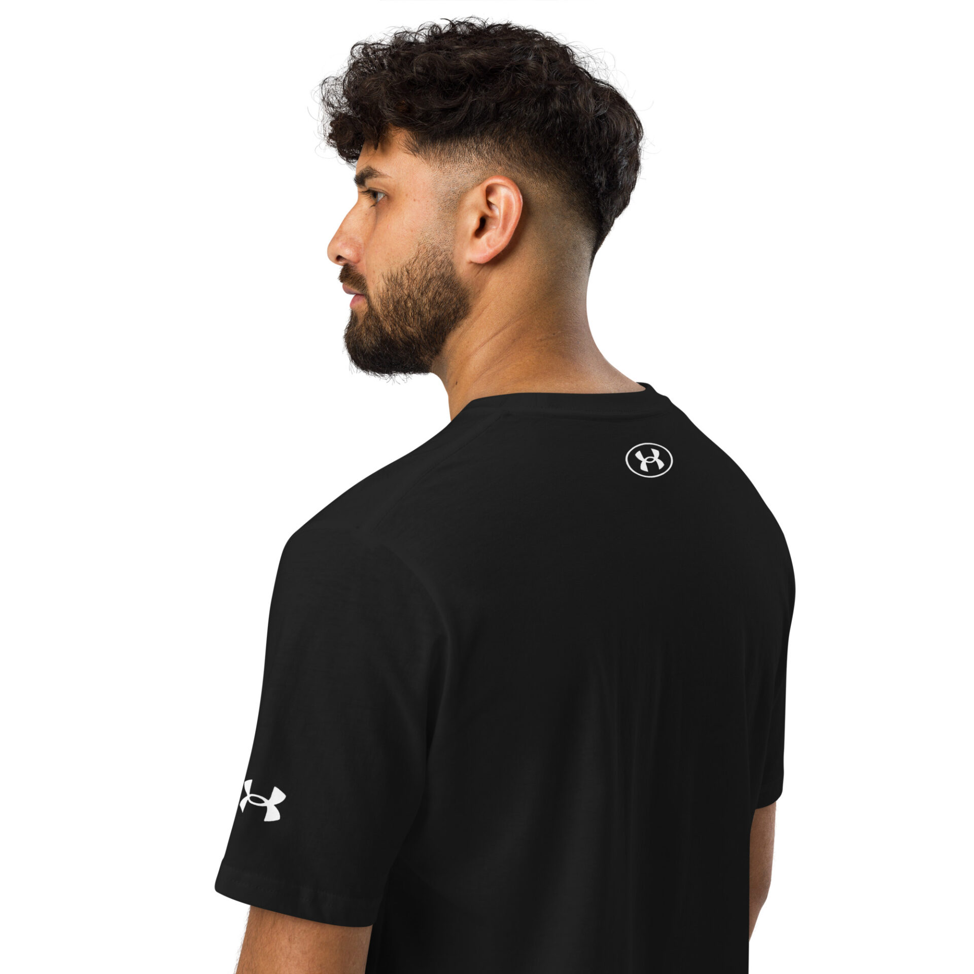 A man with dark curly hair and a beard wears the Under ArmourⓇ athletic t-shirt CHAMP in black, showing the logo on his left sleeve and upper back. He is positioned in a three-quarter view, facing away.