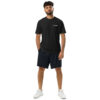 A man with curly hair and a beard stands facing forward, wearing an Under ArmourⓇ athletic t-shirt CHAMP, dark shorts, and white sneakers against a plain white background. His arms are relaxed by his sides.