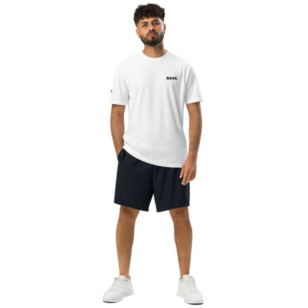 A man with curly hair and a beard stands against a white background, wearing an Under ArmourⓇ athletic t-shirt with BAAS. on the chest, black shorts, and white sneakers, hands by his sides.