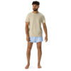 A man with curly hair and a beard stands barefoot against a white background, wearing a beige t-shirt and MJL Athletic Shorts in light blue with the MJL logo on the left leg.
