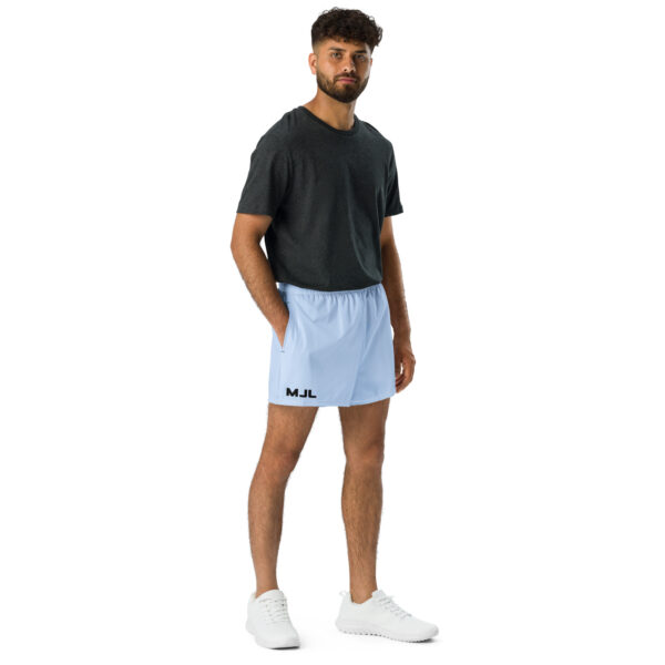 A man with short curly hair and a beard stands against a white background, wearing a dark gray T-shirt, MJL Athletic Shorts in light blue with the initials on the left leg, and white sneakers.