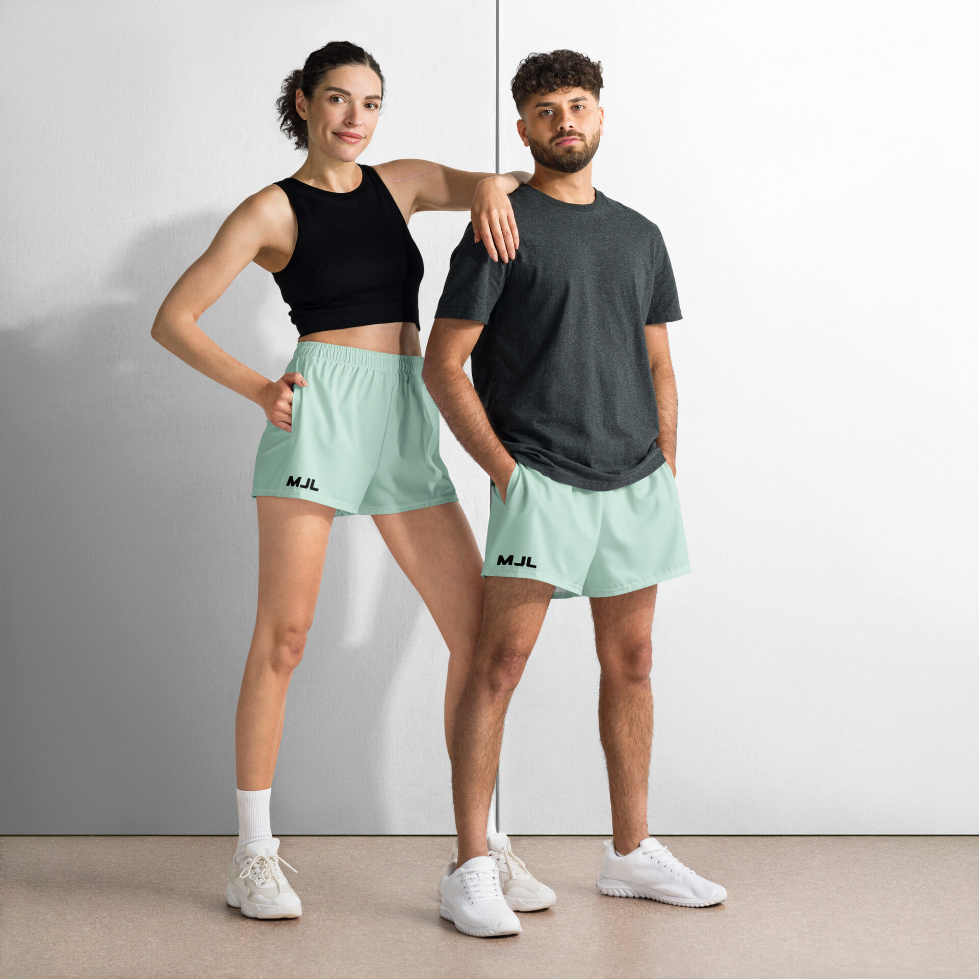 A woman and man pose confidently in mint green MJL Athletic Shorts, white sneakers, and casual tops against a plain background. The woman rests her arm on the mans shoulder, highlighting the stylish sportswear look.
