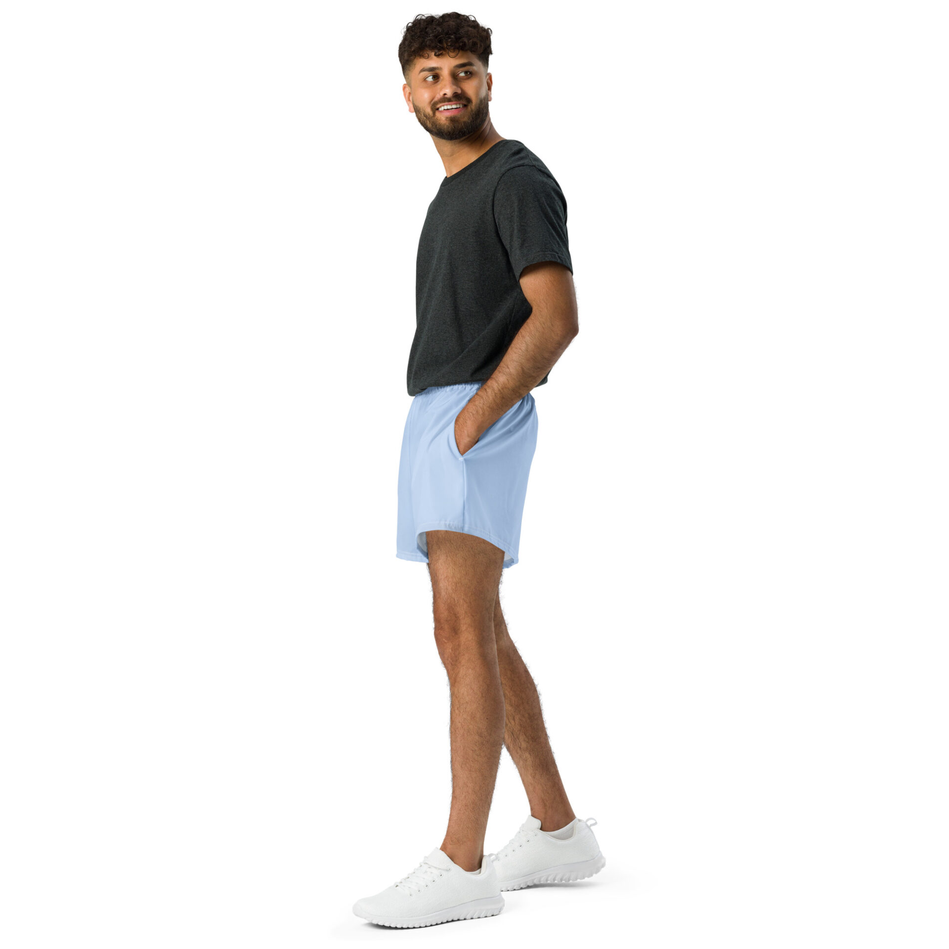 A man stands against a white background, smiling to the side. He wears MJL Athletic Shorts in light blue, paired with a dark gray t-shirt and white sneakers, his hands casually resting in the shorts pockets.
