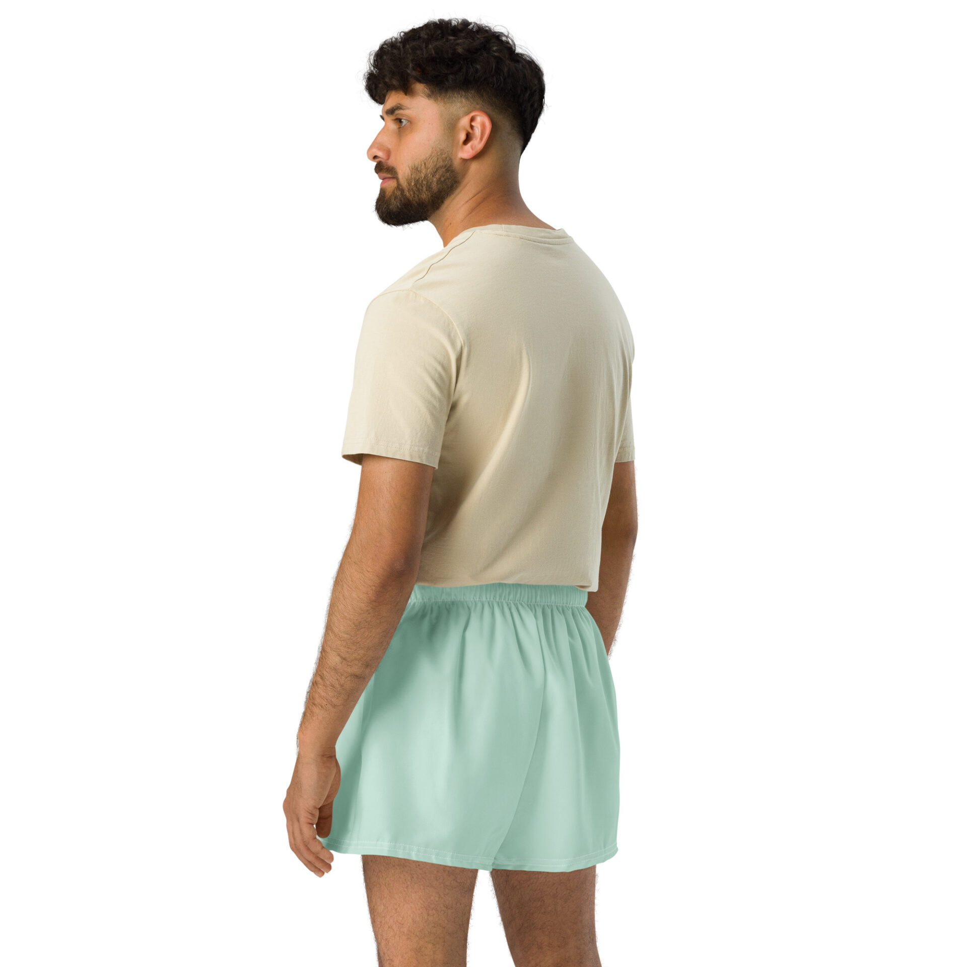 A man with dark hair and a beard wears a beige t-shirt and light green MJL Athletic Shorts, standing turned slightly away against a plain white background.