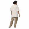 A person with long dark hair in a bun stands facing away, wearing an ADAM XXX TANTOE OUD Oversized faded t-shirt, tan cargo pants, and white shoes against a dark background.