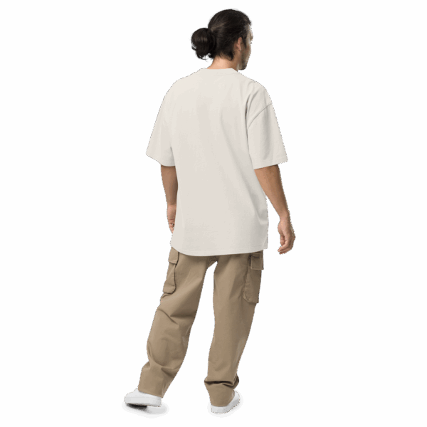 A person with long dark hair in a bun stands facing away, wearing an ADAM XXX TANTOE OUD Oversized faded t-shirt, tan cargo pants, and white shoes against a dark background.