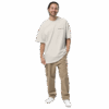 A man with long dark hair tied back is smiling and wearing an ADAM XXX TANTOE OUD Oversized faded t-shirt, khaki cargo pants, and white sneakers, standing with one hand in his pocket against a plain dark background.