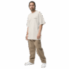 A man with long hair in a ponytail wears an ADAM XXX TANTOE OUD Oversized faded t-shirt, tan cargo pants, and white sneakers, standing against a plain black background.