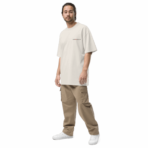 A man with long hair in a ponytail wears an ADAM XXX TANTOE OUD Oversized faded t-shirt, tan cargo pants, and white sneakers, standing against a plain black background.